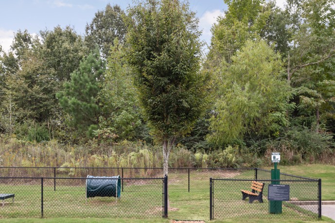 Secure gated dog park complete with a waste disposal station, agility equipment, and expansive grassy areas bordered by large trees.